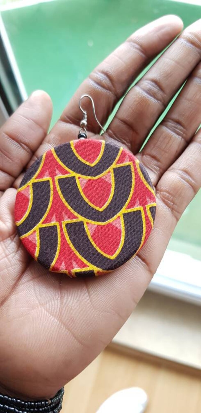 May include: A pair of large, round earrings with a geometric pattern in red, black, and yellow. The earrings are made of fabric and have a silver hook.