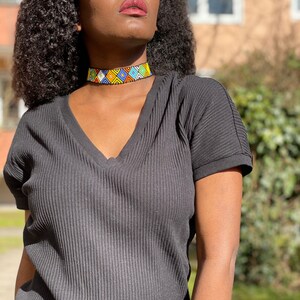 May include: A vibrant beaded choker necklace featuring a geometric design in blue, yellow, green, and white. The necklace is worn with a black V-neck top and sunglasses.