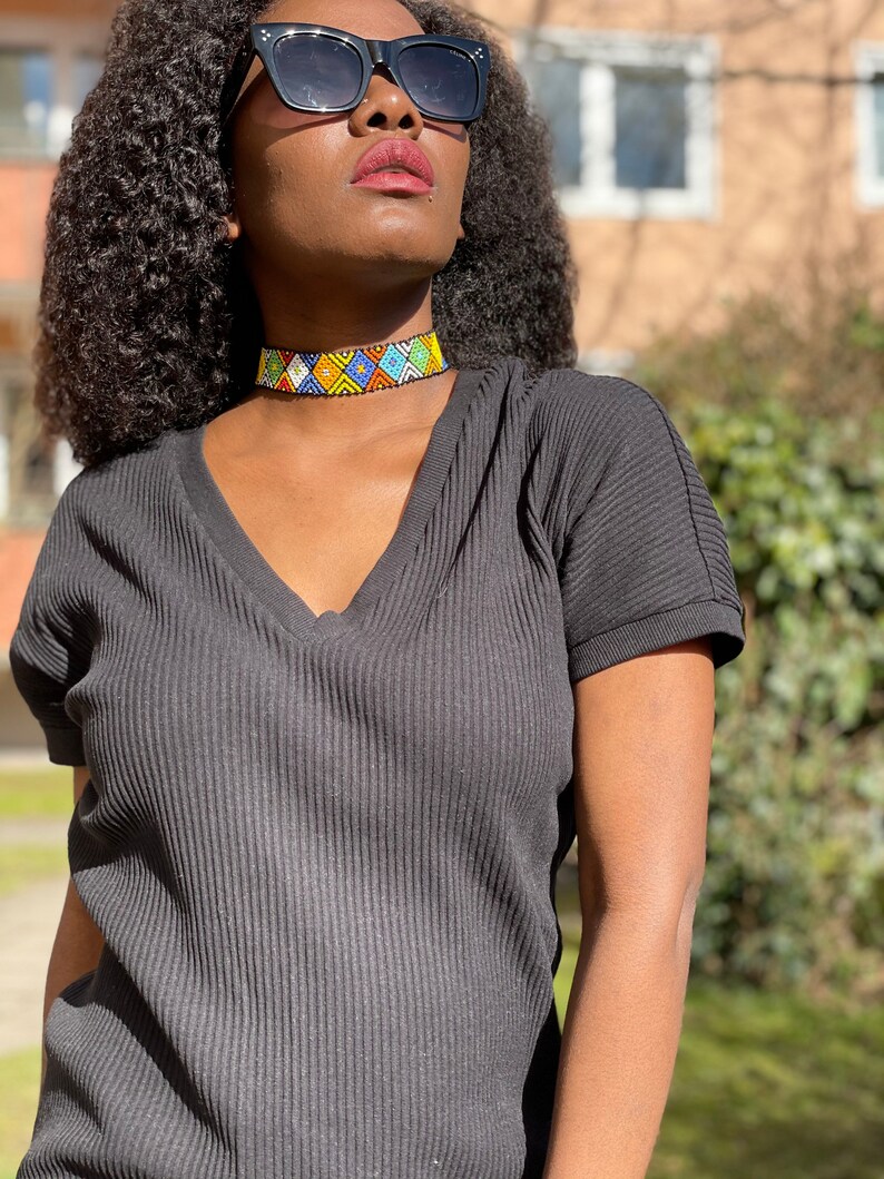 May include: A vibrant beaded choker necklace featuring a geometric design in blue, yellow, green, and white. The necklace is worn with a black V-neck top and sunglasses.