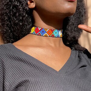 May include: A vibrant beaded choker necklace featuring a geometric design in red, blue, yellow, and white. The necklace is worn by a person in a grey V-neck top. The choker is approximately 2.5 cm wide.
