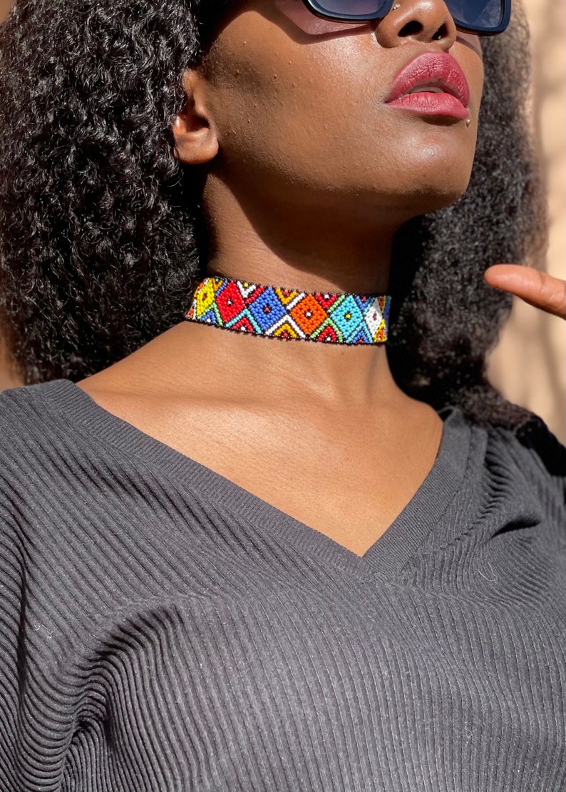 May include: A vibrant beaded choker necklace featuring a geometric design in red, blue, yellow, and white. The necklace is worn by a person in a grey V-neck top. The choker is approximately 2.5 cm wide.