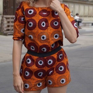 May include: An orange two-piece outfit with a repeating pattern of dark red and white circles. The top is a short-sleeved crop top, and the shorts have a black waistband. The outfit is worn by a person with curly blonde hair.