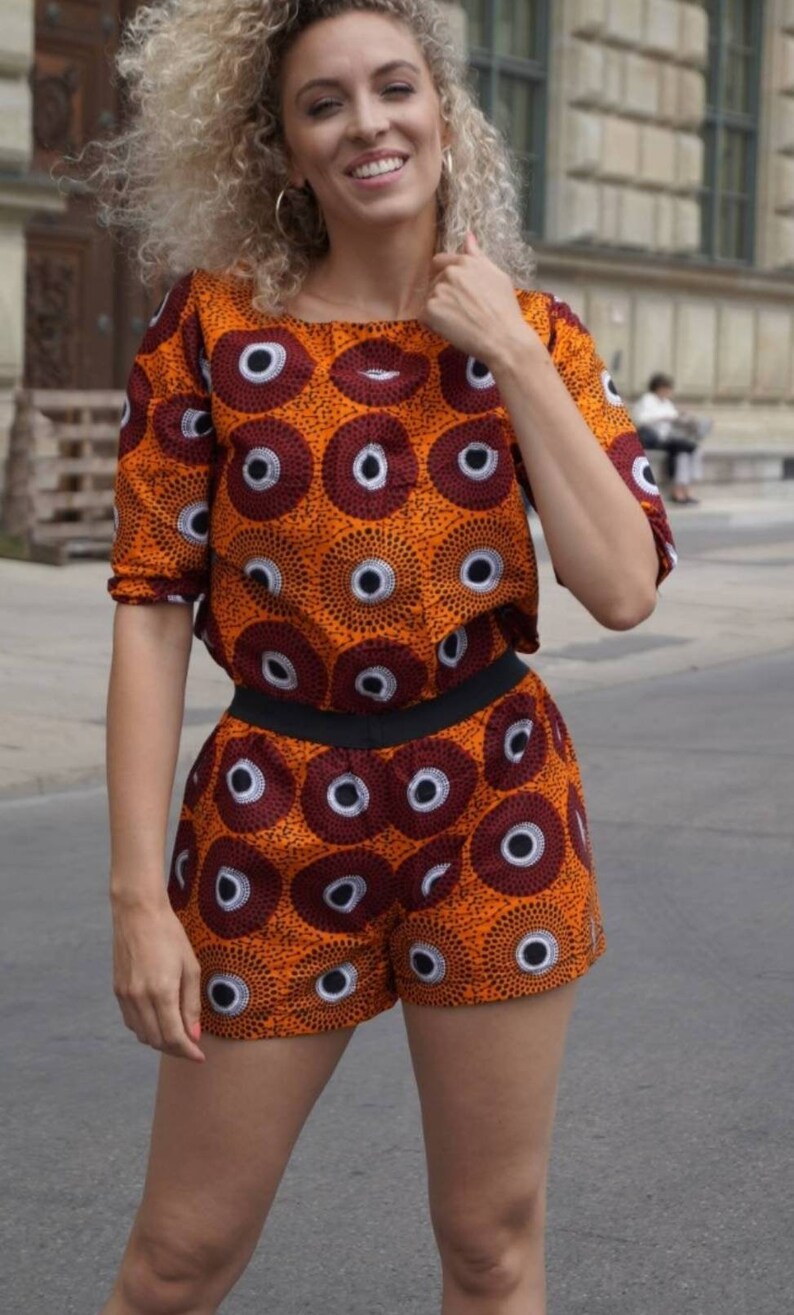 May include: An orange two-piece outfit with a repeating pattern of dark red and white circles. The top is a short-sleeved crop top, and the shorts have a black waistband. The outfit is worn by a person with curly blonde hair.