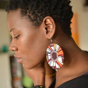 May include: A woman with short, dark hair wearing large, round earrings with a colorful geometric pattern. The earrings are brown, white, orange, and yellow.