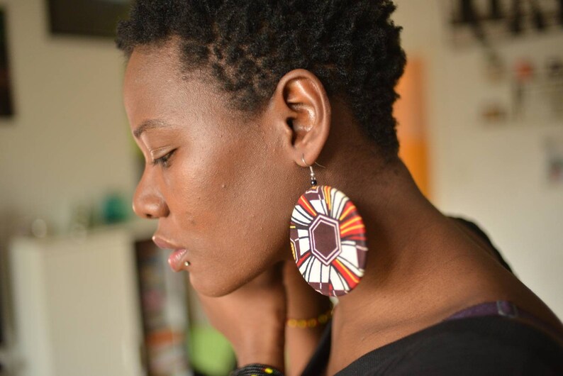 May include: A woman with short, dark hair wearing large, round earrings with a colorful geometric pattern. The earrings are brown, white, orange, and yellow.