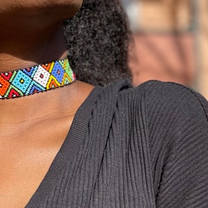 May include: A vibrant beaded choker necklace with a geometric design. The necklace showcases a mix of colours such as red, orange, blue, green, and white. The choker is worn around the neck, set against a black top.