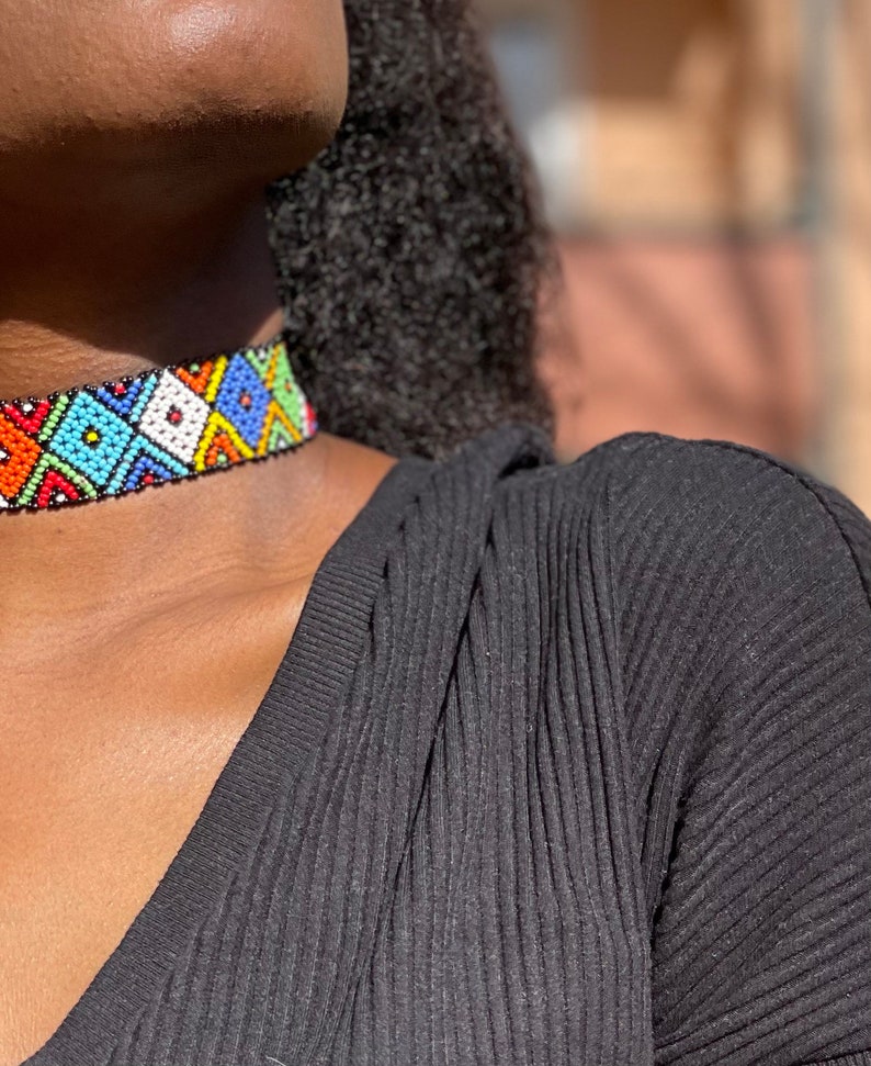 May include: A vibrant beaded choker necklace with a geometric design. The necklace showcases a mix of colours such as red, orange, blue, green, and white. The choker is worn around the neck, set against a black top.