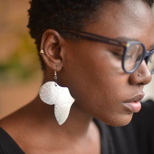 May include: A woman wearing blue-framed glasses and silver earrings shaped like the continent of Africa. The earrings have a textured, hammered finish.