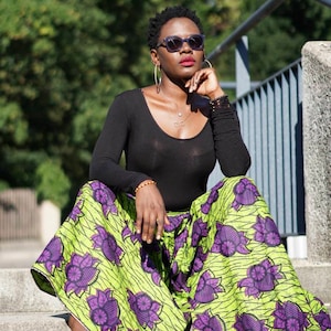May include: A woman wearing a black long-sleeved shirt and green and purple patterned palazzo pants. She is sitting on a set of concrete steps with a railing behind her. She is wearing sunglasses and has her hand on her chin.