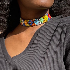 May include: A vibrant beaded choker necklace with a geometric design. The necklace showcases a black base adorned with diamond patterns in yellow, blue, orange, green, and white. The choker is worn over a black ribbed top.