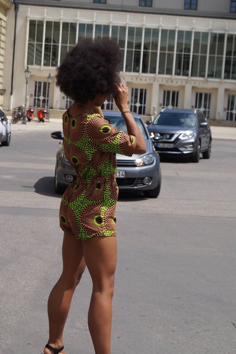 May include: A woman wearing a short-sleeved romper with a brown and green circular pattern. The romper has a scoop neckline and a fitted waist. The woman is standing on a street with cars and a building in the background.