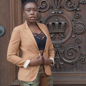 May include: A tan blazer with cream-colored cuffs and a black top. The shorts have a colorful geometric pattern. The background is a wooden door with ornate carvings.