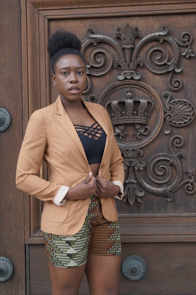 May include: A tan blazer with cream-colored cuffs and a black top. The shorts have a colorful geometric pattern. The background is a wooden door with ornate carvings.