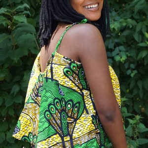 May include: A colourful sleeveless top featuring a peacock feather design in yellow, green, and blue. The top has thin straps and a flared shape. The model is wearing black trousers, with a backdrop of green foliage.