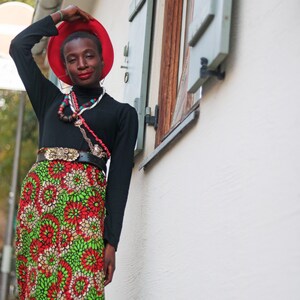 May include: A person wearing a red hat, black long-sleeved top, and a long skirt with a floral pattern in red, green, and white. They are also wearing a belt with a gold buckle and multiple necklaces.