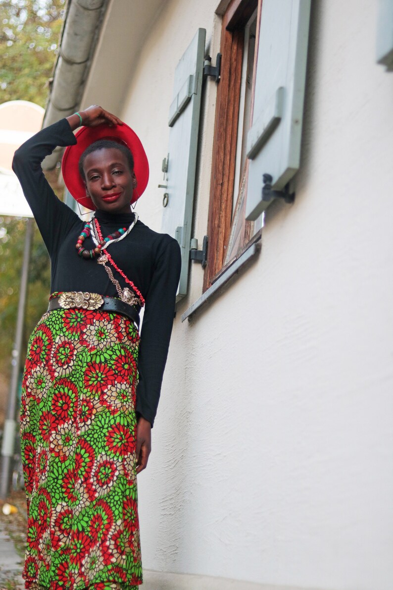May include: A person wearing a red hat, black long-sleeved top, and a long skirt with a floral pattern in red, green, and white. They are also wearing a belt with a gold buckle and multiple necklaces.