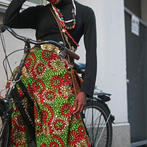 May include: A person wearing a black turtleneck, red hat, and a colourful skirt with a floral pattern. The skirt is green, red, and white. The person is leaning on a black bicycle.