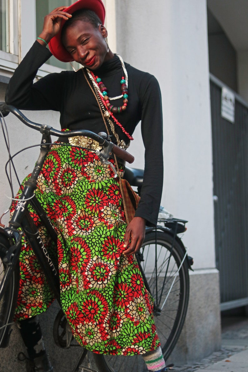 May include: A person wearing a black turtleneck, red hat, and a colourful skirt with a floral pattern. The skirt is green, red, and white. The person is leaning on a black bicycle.