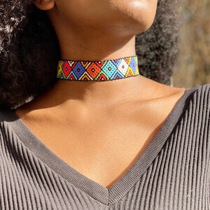 May include: A colourful beaded choker necklace. The necklace displays a geometric pattern of diamonds in red, blue, orange, yellow, and white against a black backdrop. The necklace is worn around the neck.