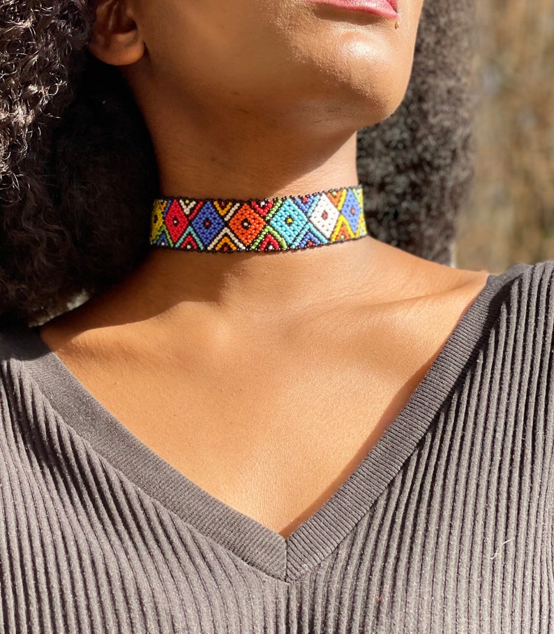 May include: A colourful beaded choker necklace. The necklace displays a geometric pattern of diamonds in red, blue, orange, yellow, and white against a black backdrop. The necklace is worn around the neck.