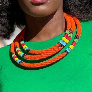 Könnte beinhalten: Eine dreireihige orangefarbene Perlenkette mit farbenfrohen Akzenten. Die Halskette hat ein lebendiges Design mit schwarzen, blauen, gelben und roten Perlen. Die Halskette wird über einem grünen Oberteil getragen.