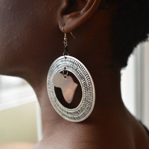 May include: Silver hoop earrings with a copper-colored cutout of the continent of Africa. The earrings are worn by a person with dark skin.