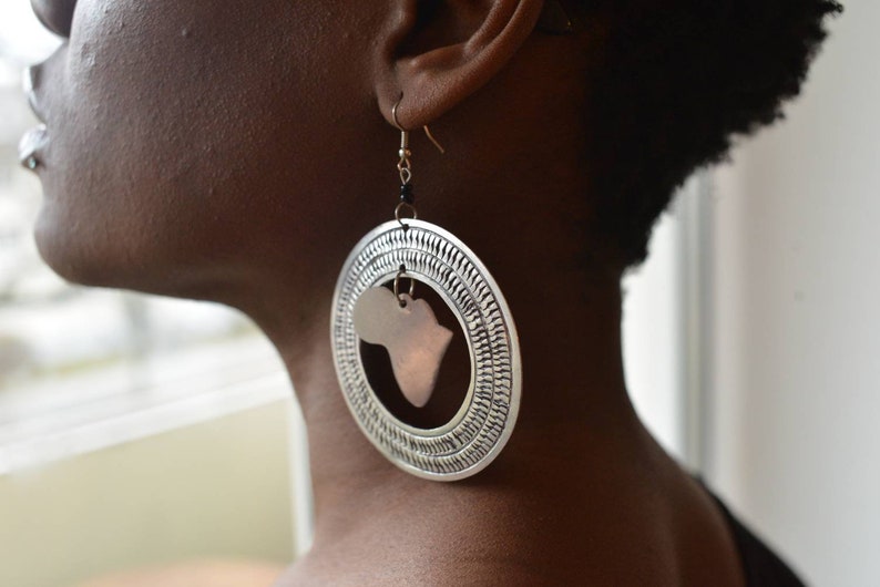 May include: Silver hoop earrings with a copper-colored cutout of the continent of Africa. The earrings are worn by a person with dark skin.