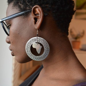May include: A close-up of a person wearing large silver earrings with a cutout of the continent of Africa. The earrings are hanging from the person's earlobes.