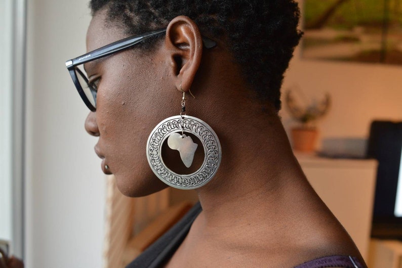 May include: A close-up of a person wearing large silver earrings with a cutout of the continent of Africa. The earrings are hanging from the person's earlobes.