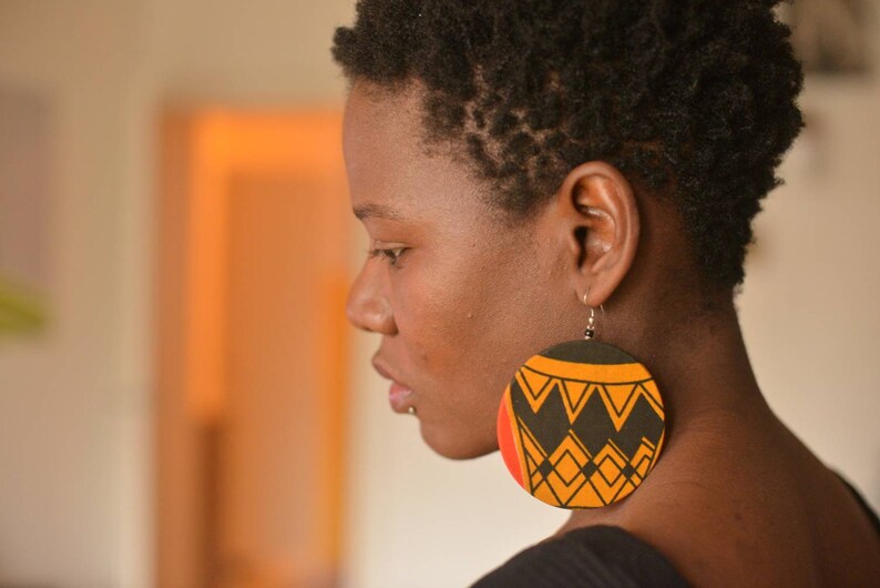 May include: A woman with short, dark hair wearing large, round earrings with a black, orange, and yellow geometric pattern.