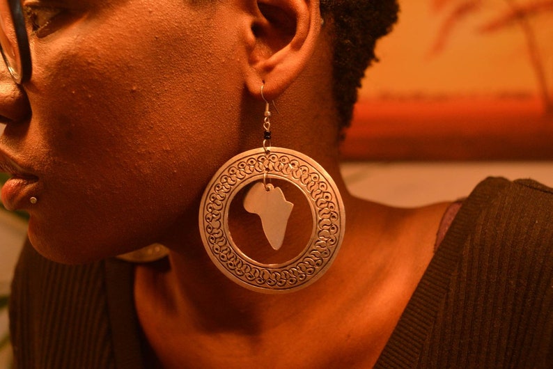 May include: Silver hoop earrings with a cutout of the continent of Africa. The earrings have a detailed, ornate design.