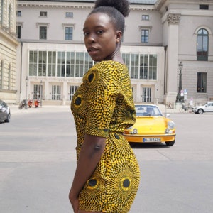 May include: A woman wearing a yellow and black patterned romper with a circular design. The romper has short sleeves and a matching shorts. A yellow vintage car is parked in the background.