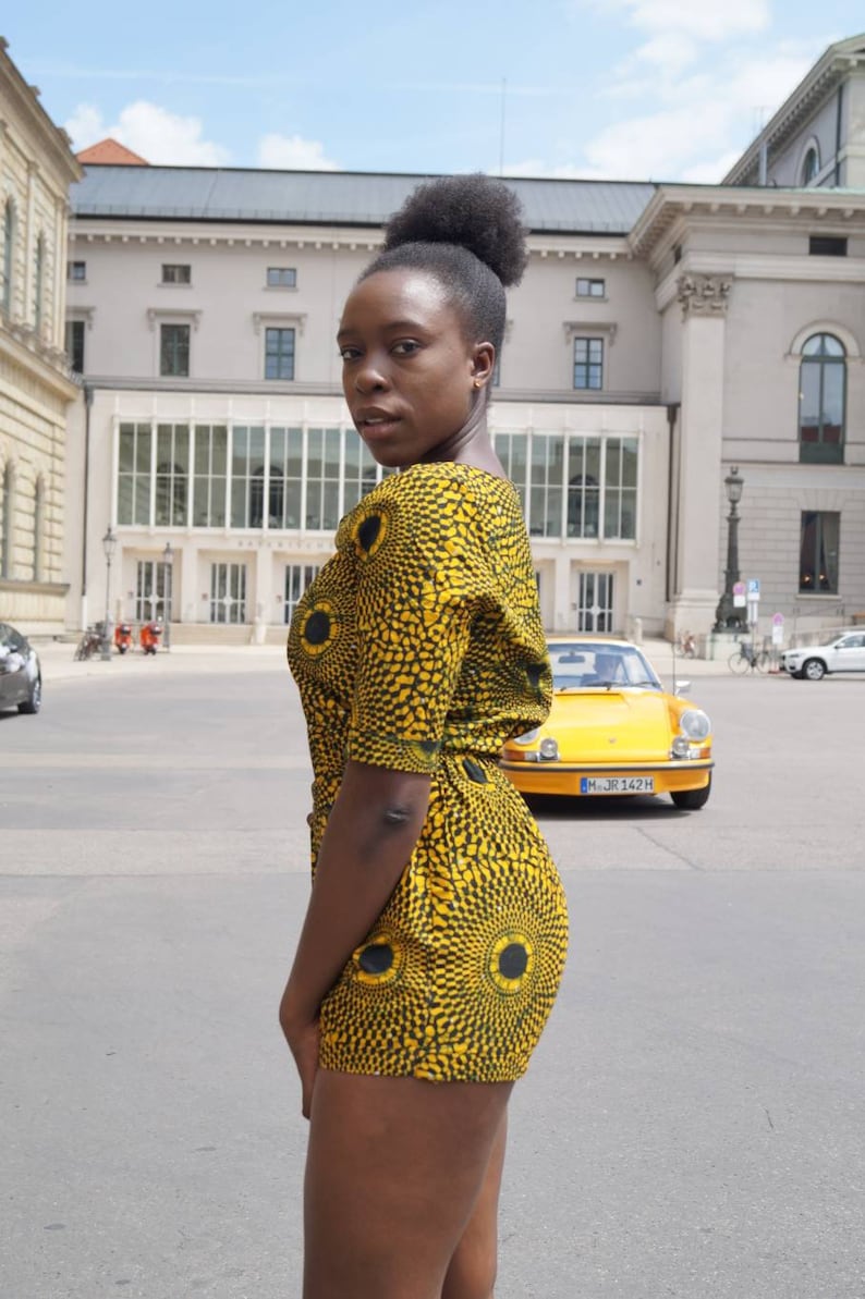 May include: A woman wearing a yellow and black patterned romper with a circular design. The romper has short sleeves and a matching shorts. A yellow vintage car is parked in the background.