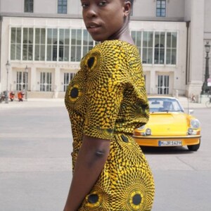 May include: A woman wearing a yellow and black patterned romper with a matching design. The romper features a circular pattern. A yellow vintage car is parked in the background.