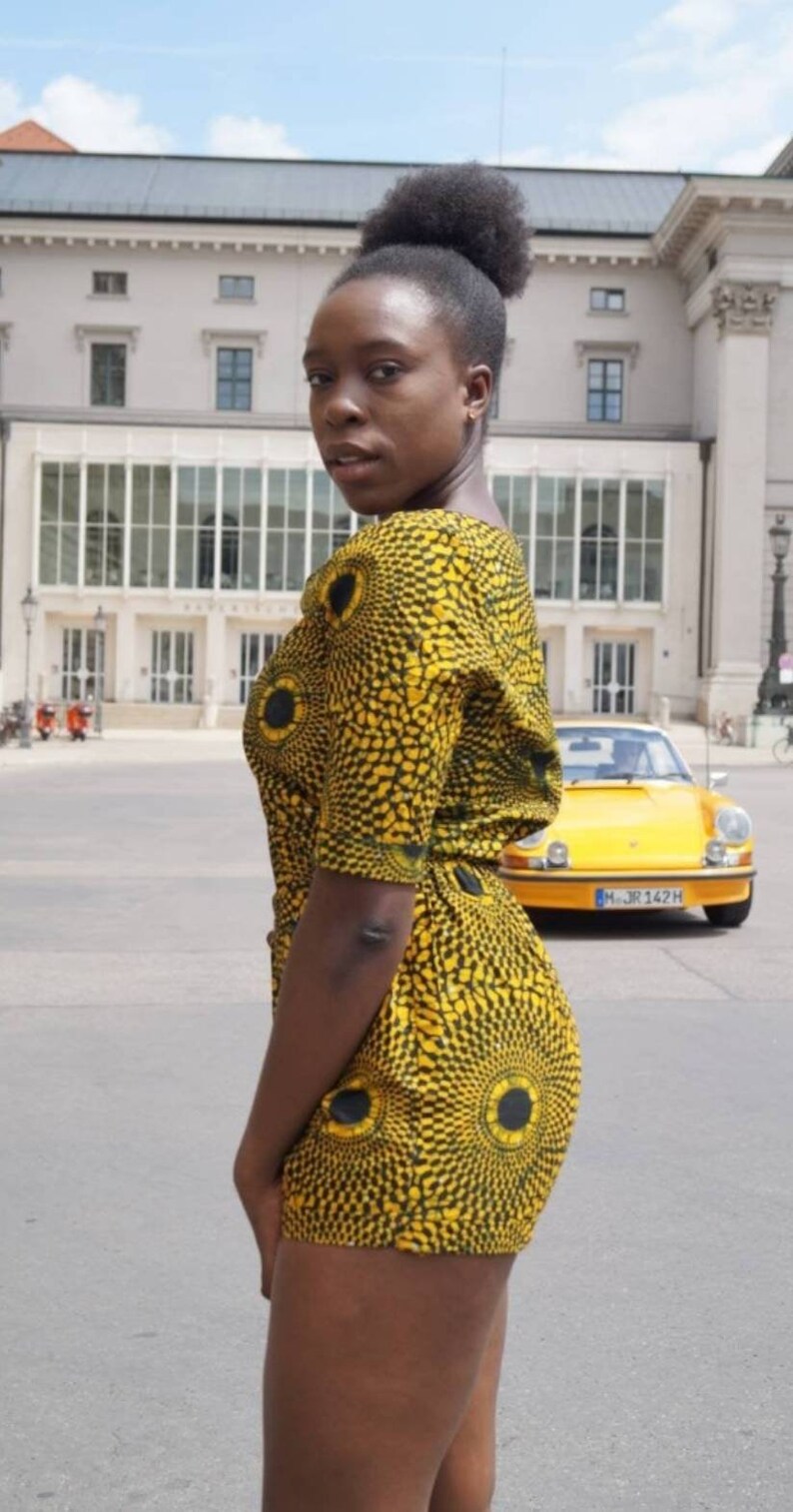 May include: A woman wearing a yellow and black patterned romper with a matching design. The romper features a circular pattern. A yellow vintage car is parked in the background.