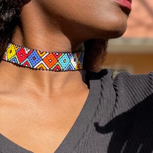 May include: A vibrant beaded choker necklace with a geometric design. The necklace displays a black base adorned with colourful beads in red, blue, yellow, orange, and white. The choker is worn around the neck, highlighting the detailed beadwork.