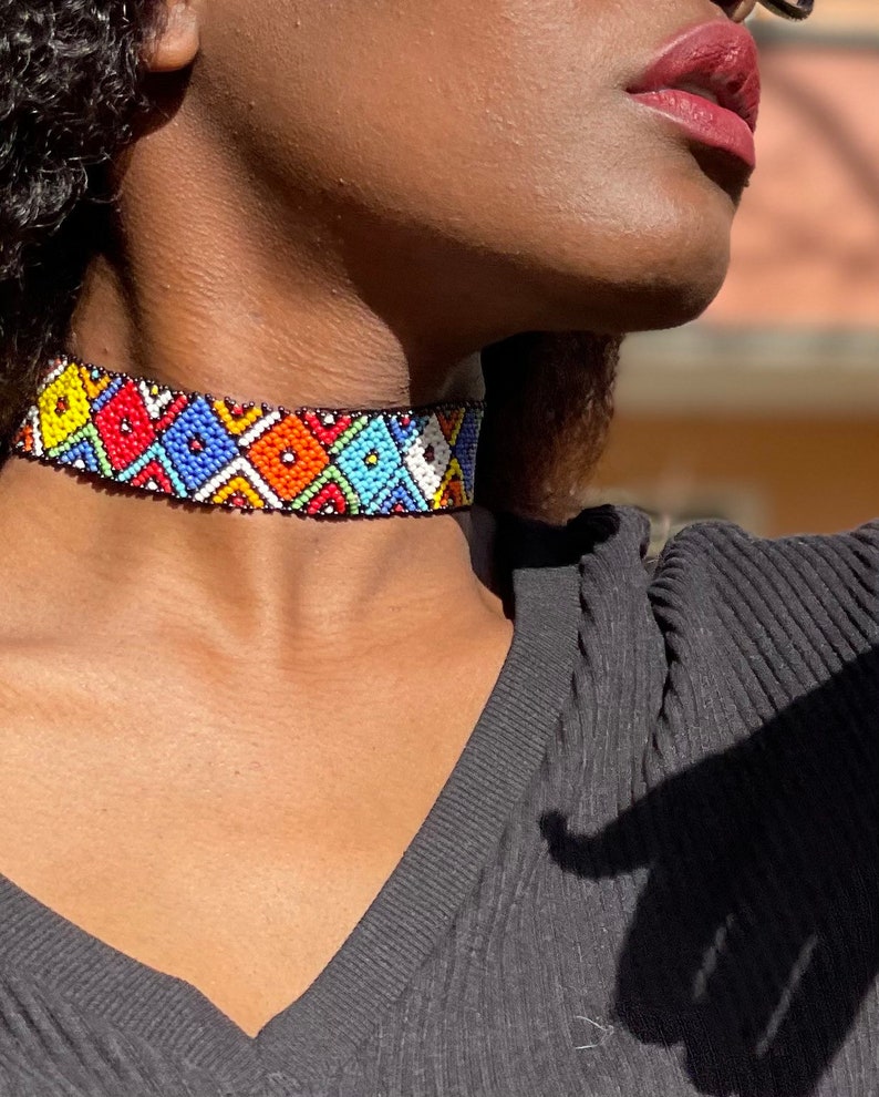 May include: A vibrant beaded choker necklace with a geometric design. The necklace displays a black base adorned with colourful beads in red, blue, yellow, orange, and white. The choker is worn around the neck, highlighting the detailed beadwork.