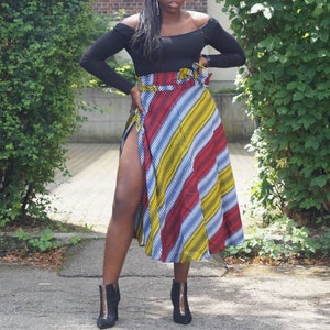 May include: A woman in a black off-the-shoulder top and a midi skirt with red, yellow, and blue diagonal stripes. She is wearing black ankle boots. The skirt has a high slit.