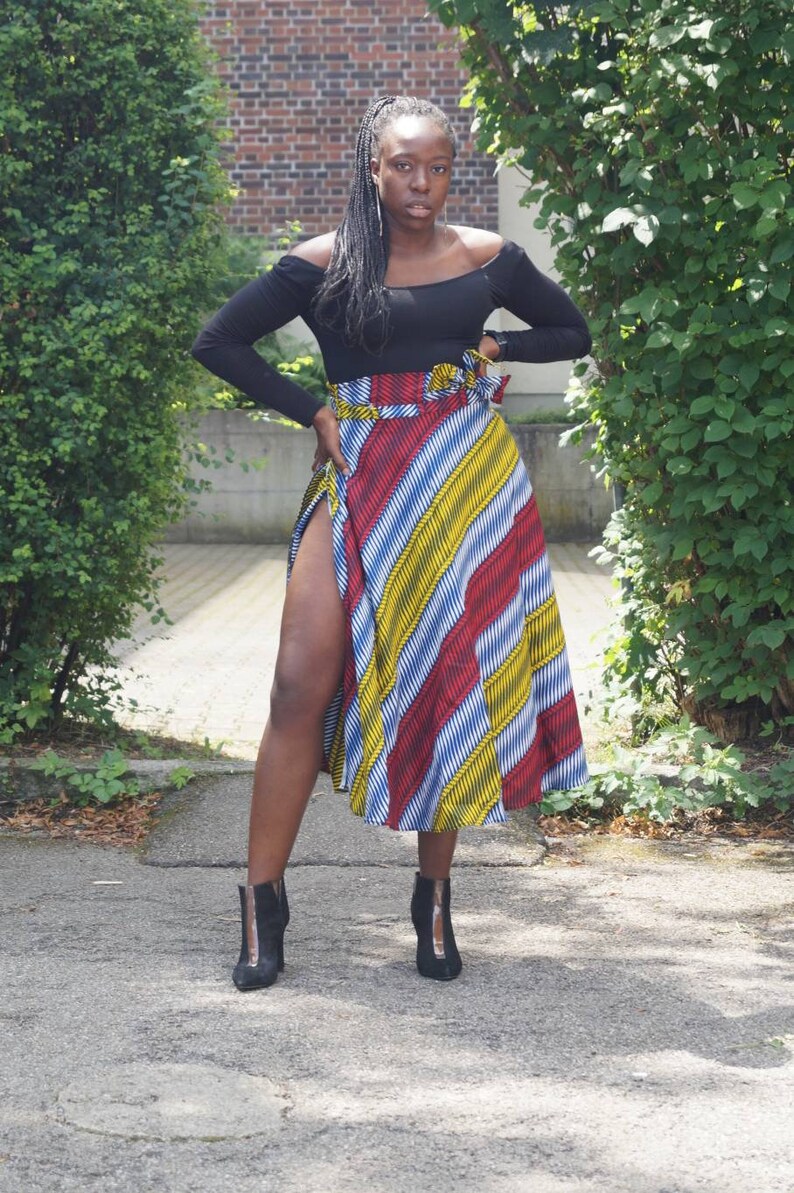 May include: A woman in a black off-the-shoulder top and a midi skirt with red, yellow, and blue diagonal stripes. She is wearing black ankle boots. The skirt has a high slit.