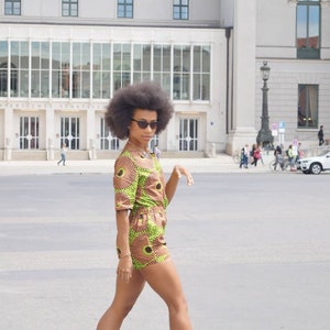 May include: A person wearing a green, brown, and yellow patterned romper with black heeled sandals and sunglasses. The outfit has a circular design. The person is walking on a paved surface in front of a large building.
