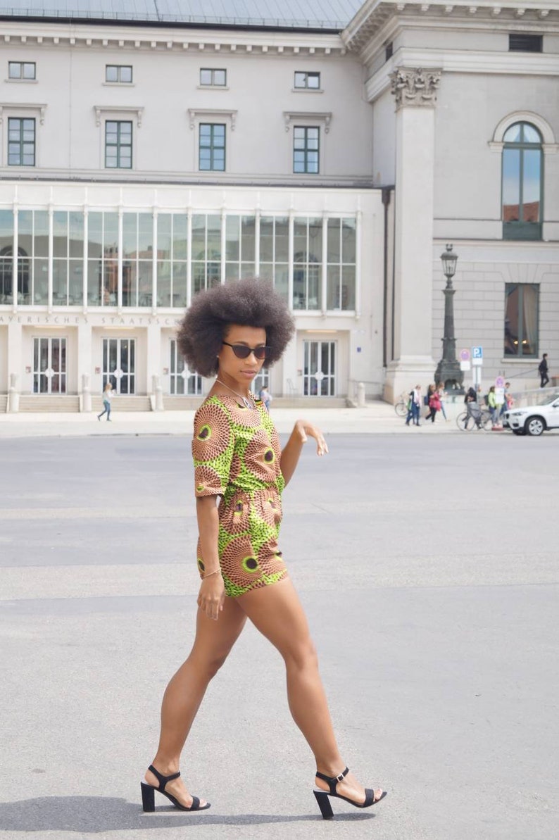 May include: A person wearing a green, brown, and yellow patterned romper with black heeled sandals and sunglasses. The outfit has a circular design. The person is walking on a paved surface in front of a large building.