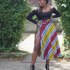 May include: A black off-the-shoulder long-sleeve top and a colourful, patterned skirt with red, yellow, and blue stripes. The skirt has a high slit. Black ankle boots complete the outfit.
