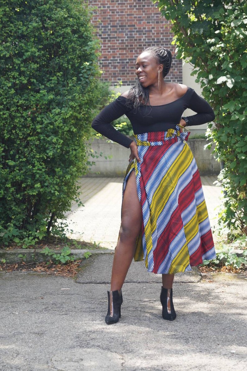 May include: A black off-the-shoulder long-sleeve top and a colourful, patterned skirt with red, yellow, and blue stripes. The skirt has a high slit. Black ankle boots complete the outfit.