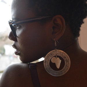 May include: A woman wearing glasses and a black top with large silver earrings shaped like the continent of Africa.