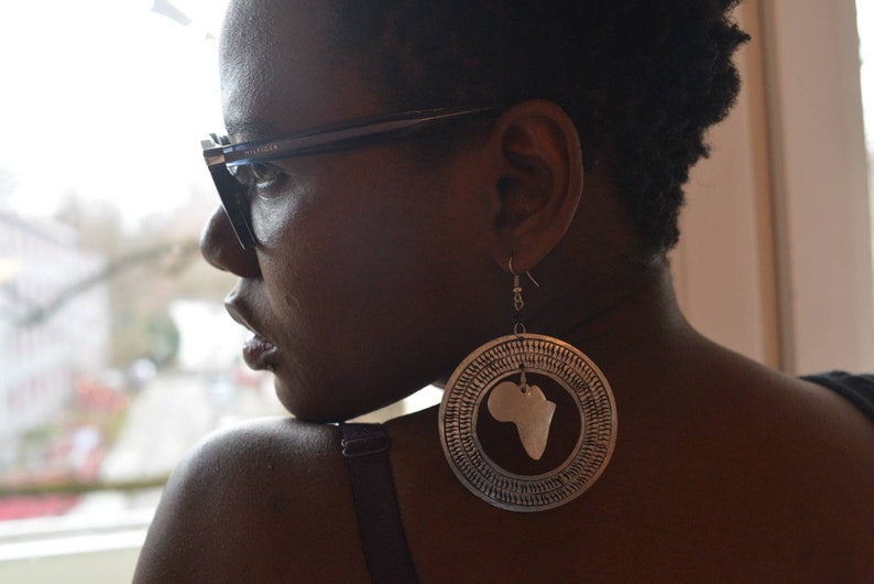 May include: A woman wearing glasses and a black top with large silver earrings shaped like the continent of Africa.