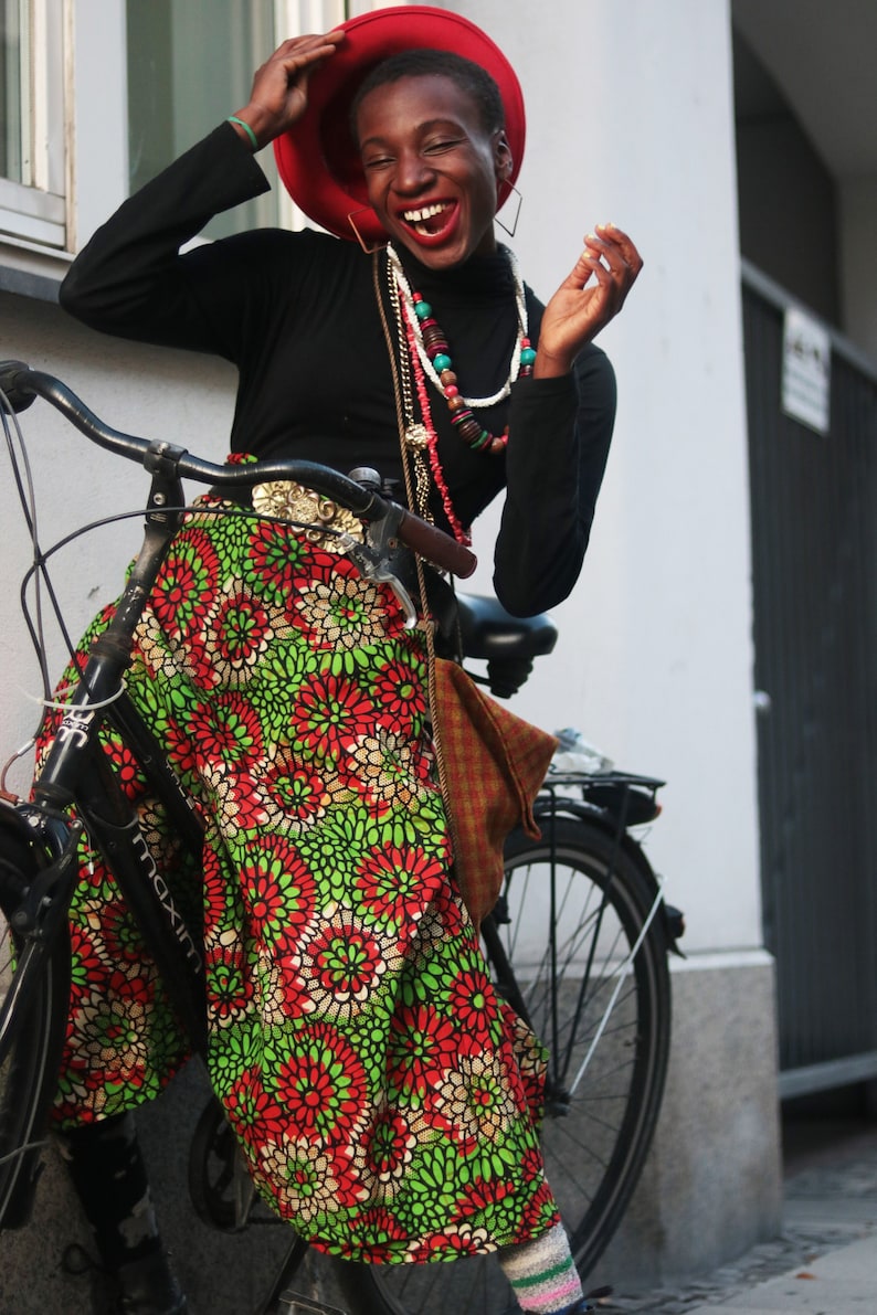 May include: A black bicycle with a woman wearing a black turtleneck and a red hat. She is wearing a colourful skirt with a floral pattern in red, green, and white. The woman is smiling and wearing multiple necklaces.