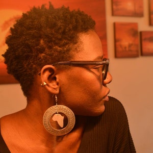 May include: A woman with short, curly brown hair wearing black framed glasses and a black shirt. She is wearing large silver earrings with a cutout of the continent of Africa.