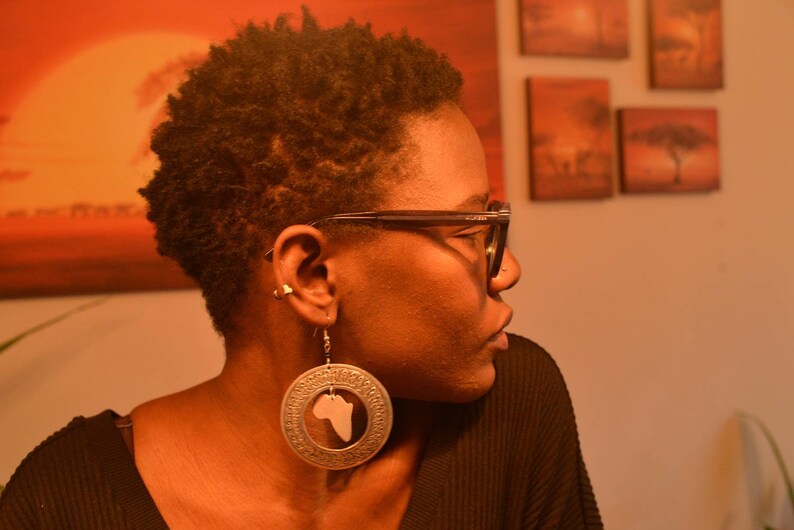 May include: A woman with short, curly brown hair wearing black framed glasses and a black shirt. She is wearing large silver earrings with a cutout of the continent of Africa.