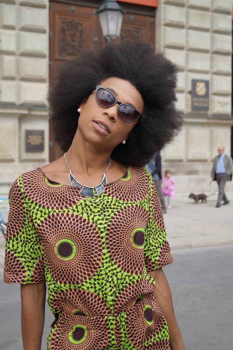 May include: A woman wearing a short-sleeved dress with a brown and green circular pattern. She is wearing black sunglasses and a silver necklace. Her hair is large and dark. The background includes a building and people.