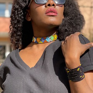 May include: A beaded choker necklace and bracelet set. The choker features a geometric pattern in blue, yellow, white, and orange beads. The bracelet is black with yellow and red accents. The person is wearing a black V-neck top.